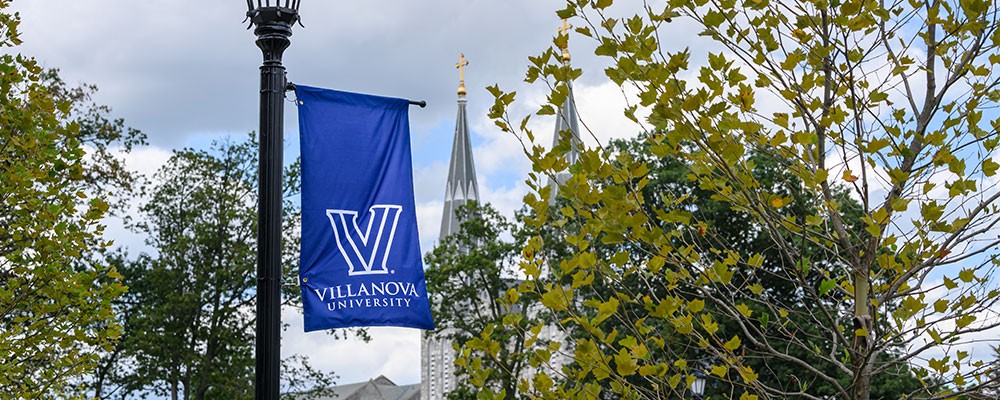 A View of the Villanova "V" logo on a blue flag banner with St. Thomas of Villanova Church in the distance